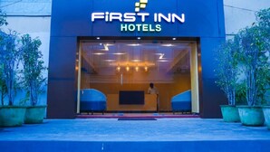 The exterior of First Inn Hotels with a modern design, featuring a large glass entrance and potted plants on either side.