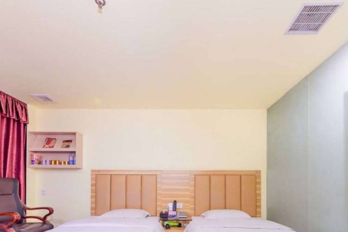 A hotel room with two single beds, a wooden headboard, a small table with a lamp, and a shelf with books.
