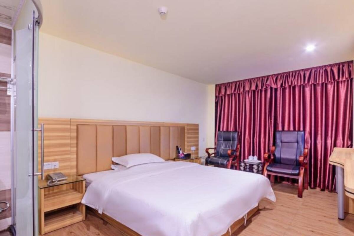 A hotel room with a large bed, wooden headboard, a desk with a chair, a small table with a lamp, and a red curtain.