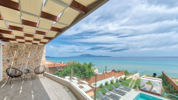 Un balcón con muro de piedra, dos sillas, una mesita, una piscina y vistas al mar y las montañas.