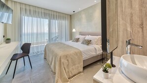 Un dormitorio moderno con una cama grande, una mesita de noche, una silla, un ventanal con cortinas translúcidas, vistas al mar, y un baño con lavamanos y espejo amplios.