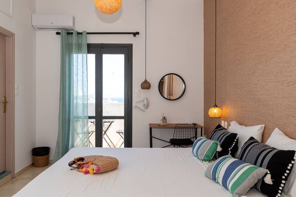 A bedroom with a bed, pillows, a bedside table, a mirror, a hanging light, and a view of the outdoors.