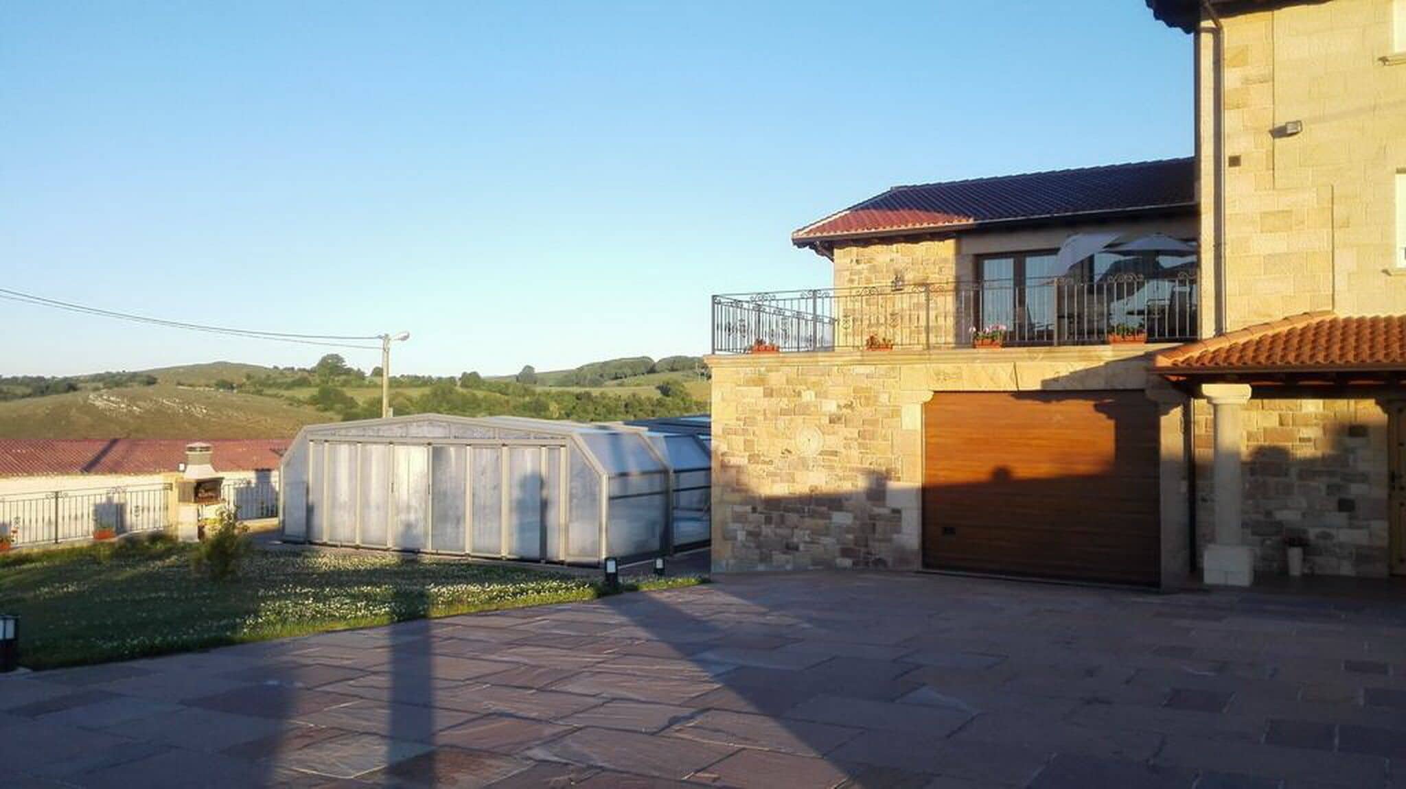 Une maison avec une façade en pierre, un balcon doté d’une balustrade en métal et une grande serre située dans le jardin arrière.