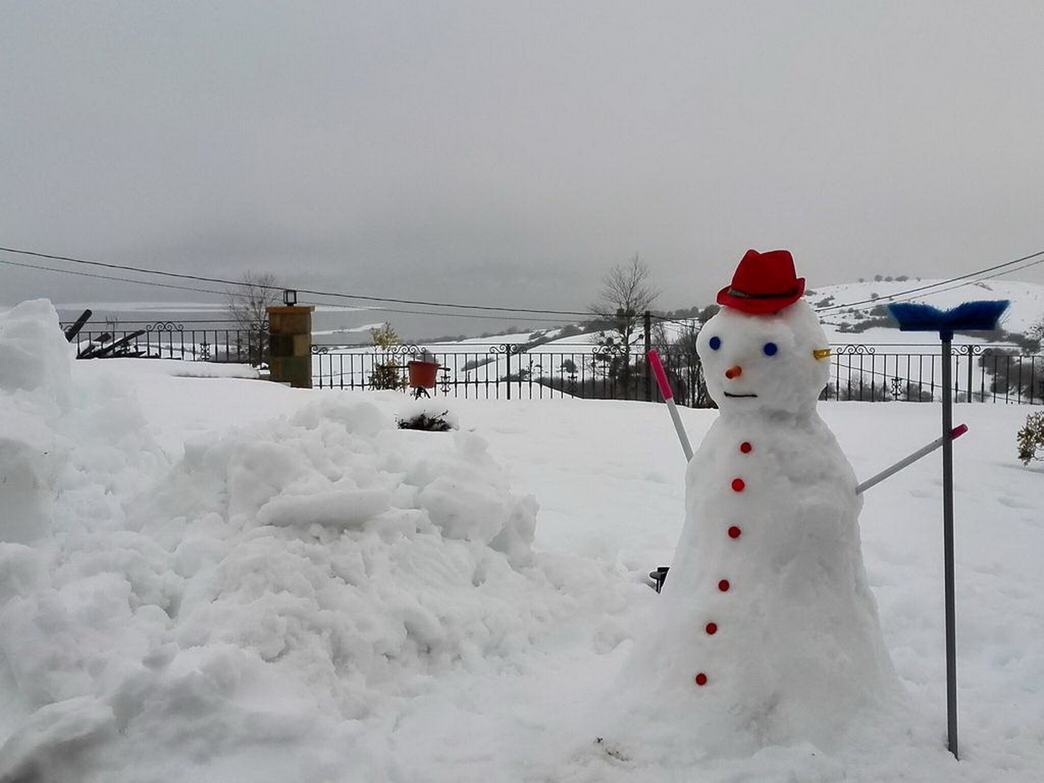 Un muñeco de nieve con sombrero rojo y botones, parado junto a una cerca en un paisaje nevado.
