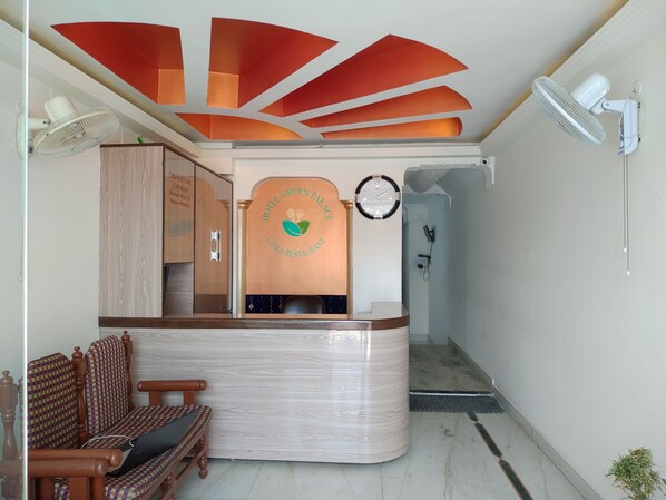 A hotel reception area with a wooden reception desk, a clock, a wall-mounted fan, and a bench.