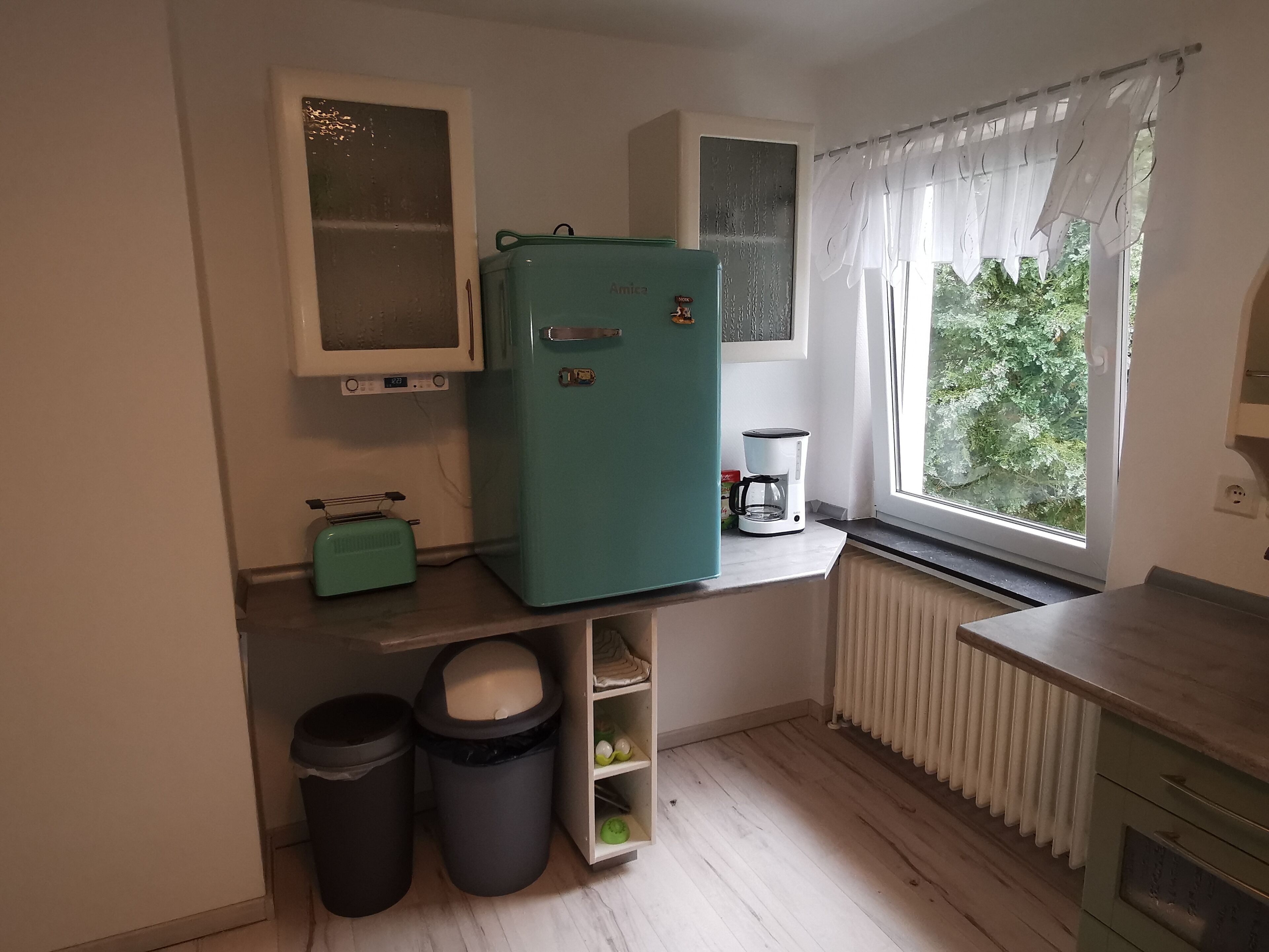 Cocina compacta con un refrigerador verde azulado, microondas, cafetera y ventana con vistas a la vegetación. Hay dos cubetas de basura y un estante con utensilios de cocina.
