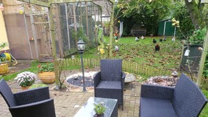 Une terrasse dotée de meubles en osier et d’une table, entourée d’un jardin où l’on peut voir des poules et un abri de jardin vert.