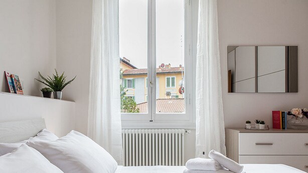 A hotel room with a bed, a window with curtains, a radiator, a bedside table with books and a vase, and a view of buildings outside. - San Frediano Apartment (Florence)