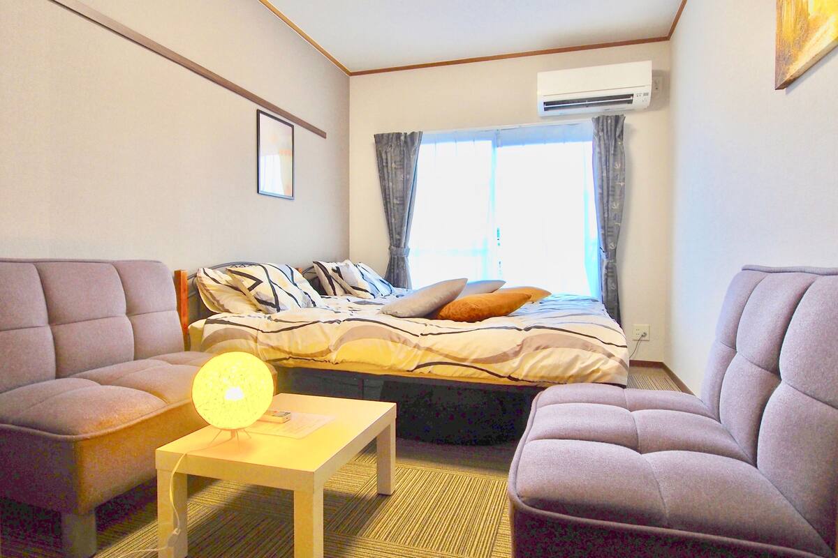 A modern hotel room with a bed, two armchairs, a small table with a lamp, and an air conditioning unit.