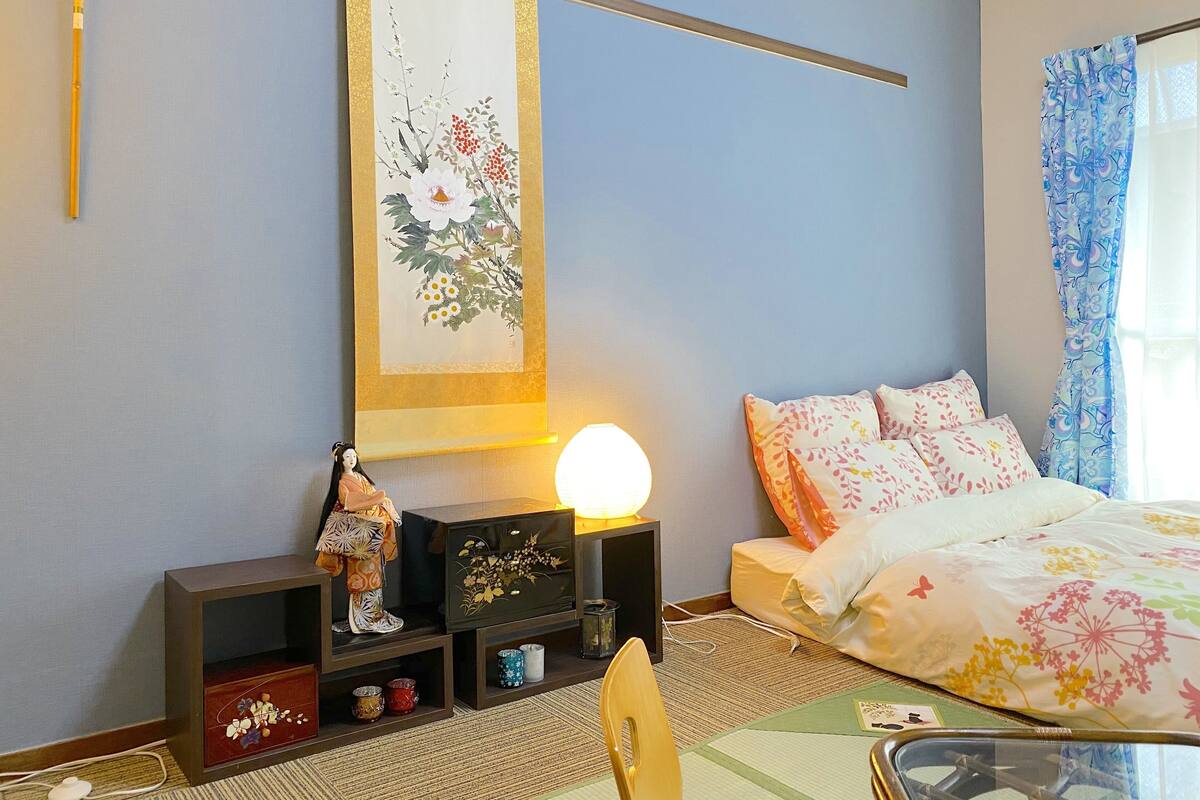 A bedroom with a bed, a nightstand, a lamp, a shelf with decorative items, and a hanging floral artwork.
