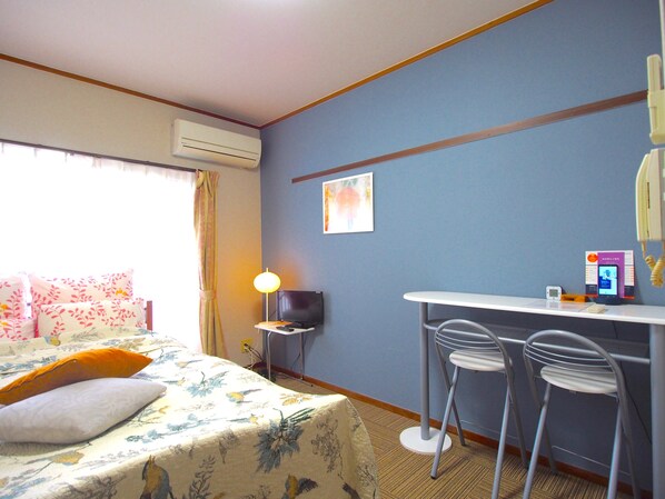 A hotel room with a bed, a desk with two chairs, an air conditioner, a lamp, and a wall-mounted phone.