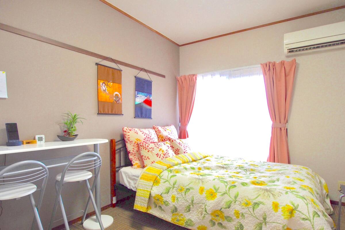 A bedroom with a bed, a desk, and two chairs. There is a window with pink curtains, an air conditioner, and a wall-mounted artwork.