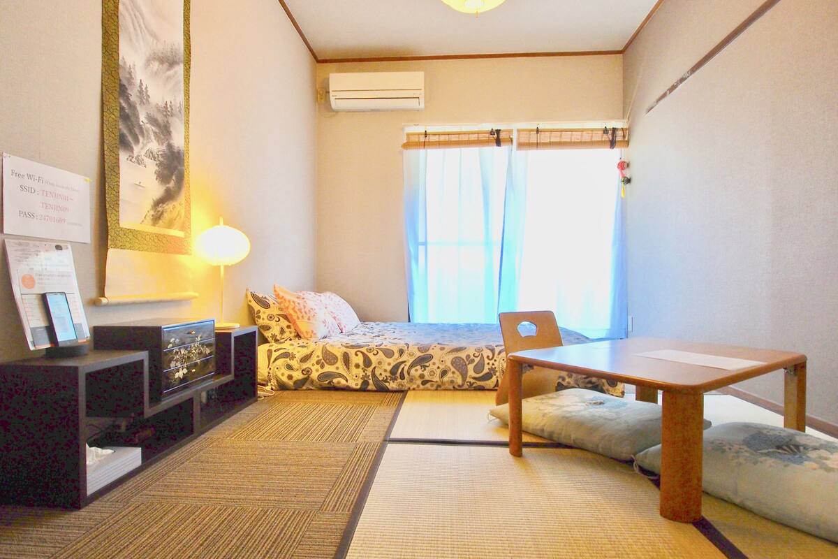 A small, neatly arranged room with a bed, a desk, a chair, and a lamp. There is a window with curtains, an air conditioning unit, and a wall-mounted shelf with a lamp and a framed picture.