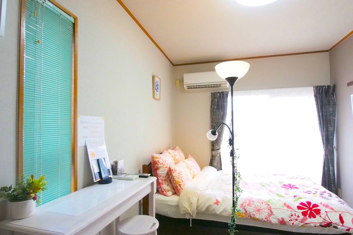 A hotel room with a bed, a desk with a chair, a lamp, a window with curtains, and a wall-mounted air conditioner.