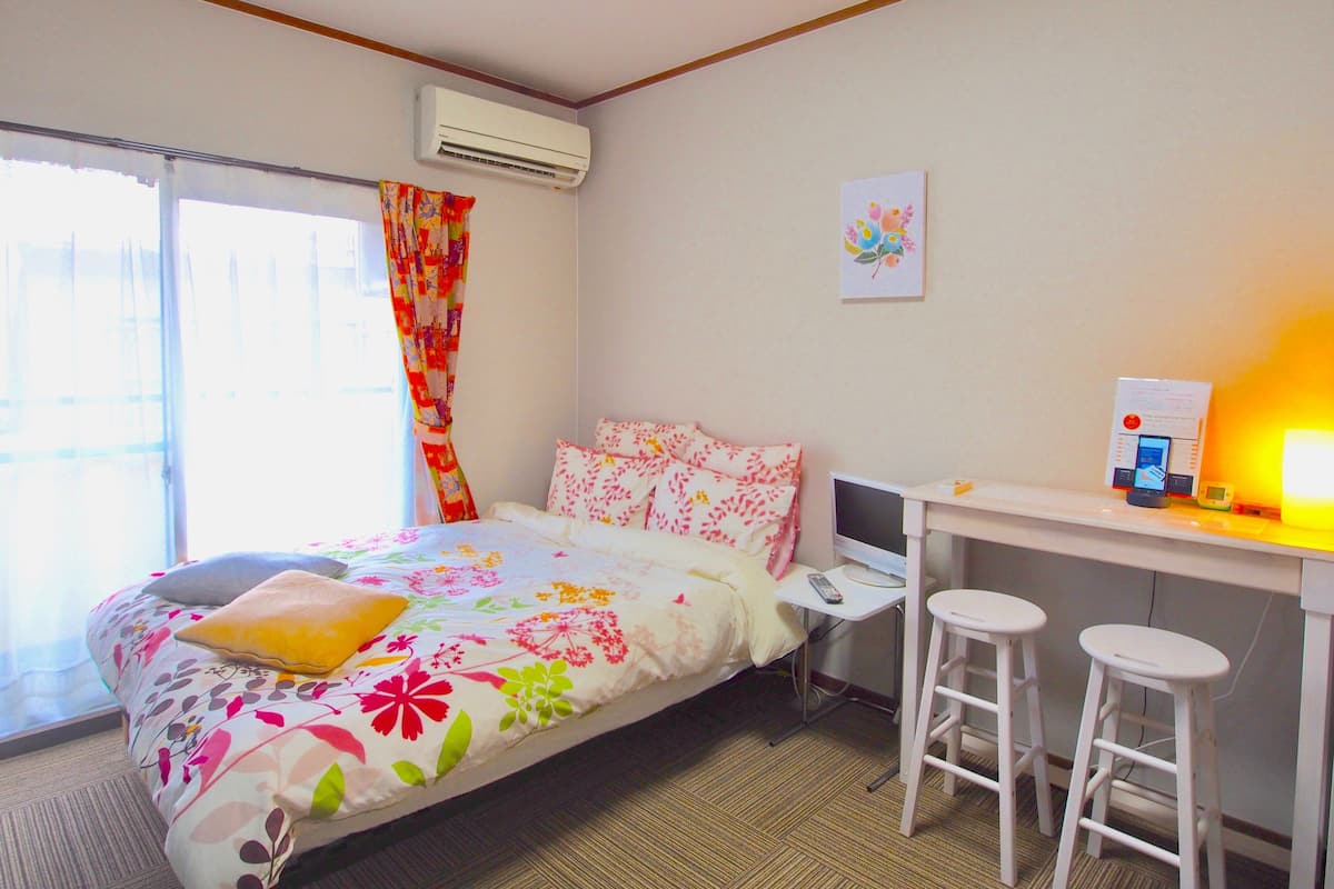 A hotel room with a bed, a desk with two stools, an air conditioner, and a window with curtains.