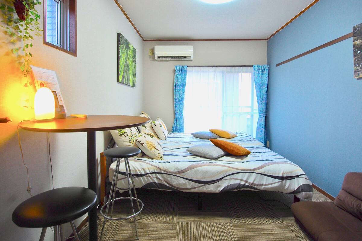 A small, well-lit room with a bed, a desk, a chair, and a hanging plant.