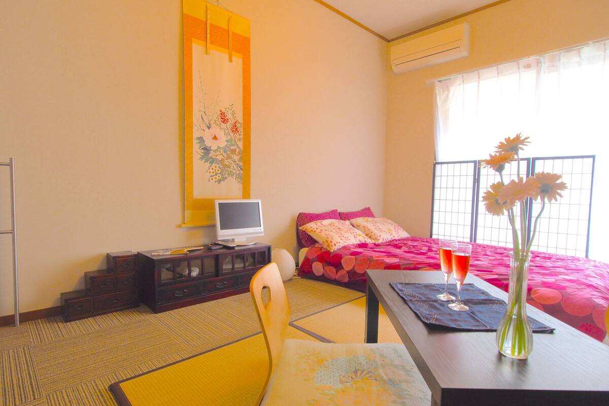 A hotel room with a bed, a desk with a computer, a chair, a table with a vase of flowers, and a window with blinds.