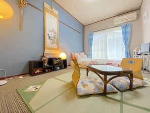 A traditional Japanese-style room with tatami flooring, a low table with cushions, a hanging scroll, and a bed with a floral bedspread.