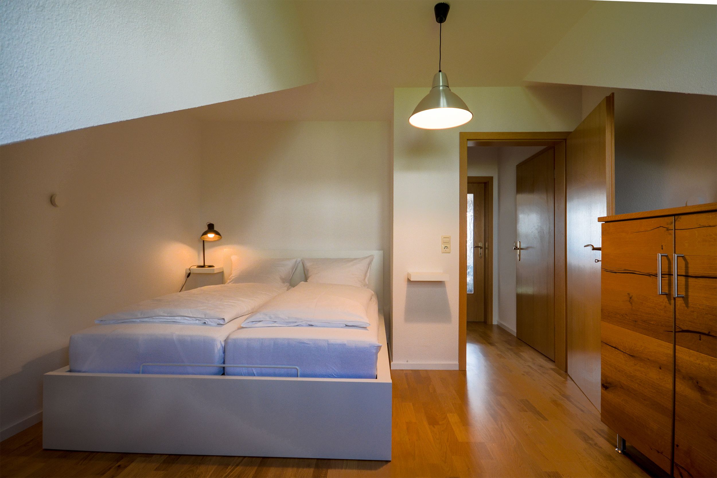 A bedroom with a bed, bedside lamp, wooden wardrobe, and a hallway leading to another room.