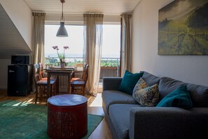 A living room with a sofa, a dining table, and a red stool. There is a painting on the wall and a view of the outdoors through the windows. - Das Nest am Wohrenberg (Daisendorf)