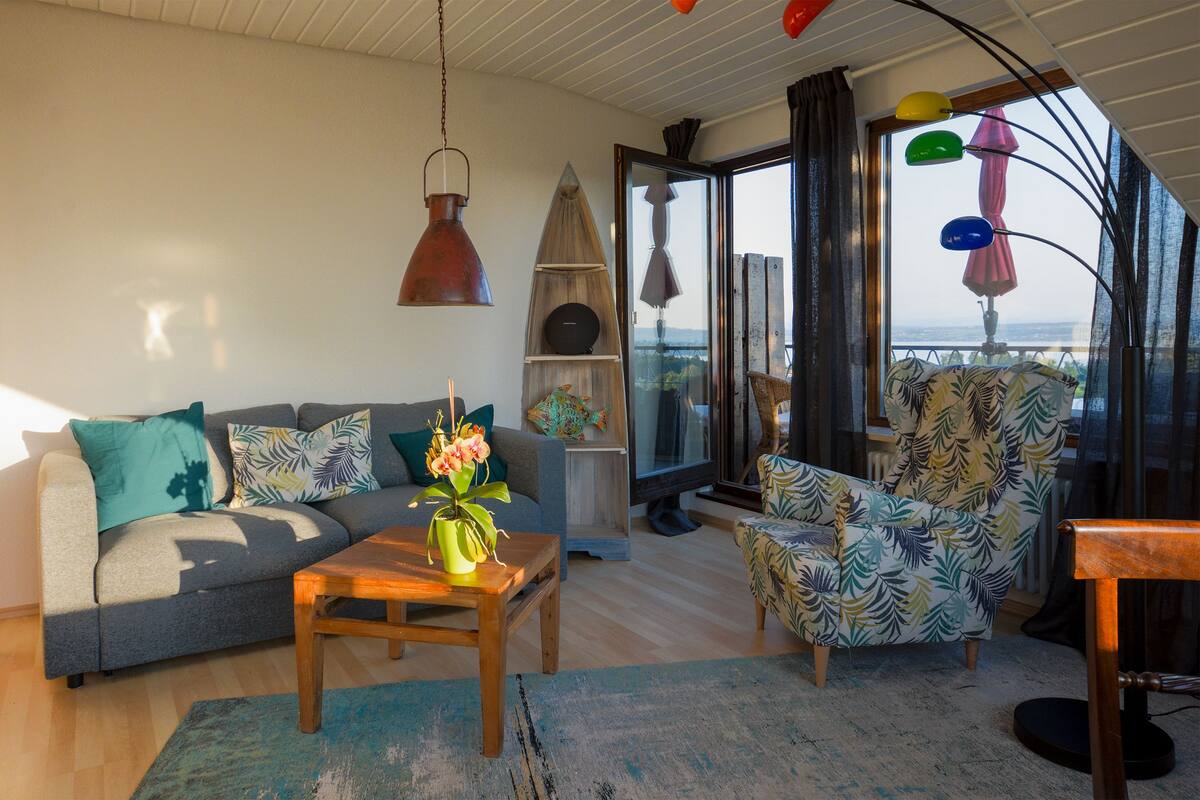 Uma sala de estar moderna com sofá, poltrona e mesa de centro. Há uma janela grande com vista para o exterior.