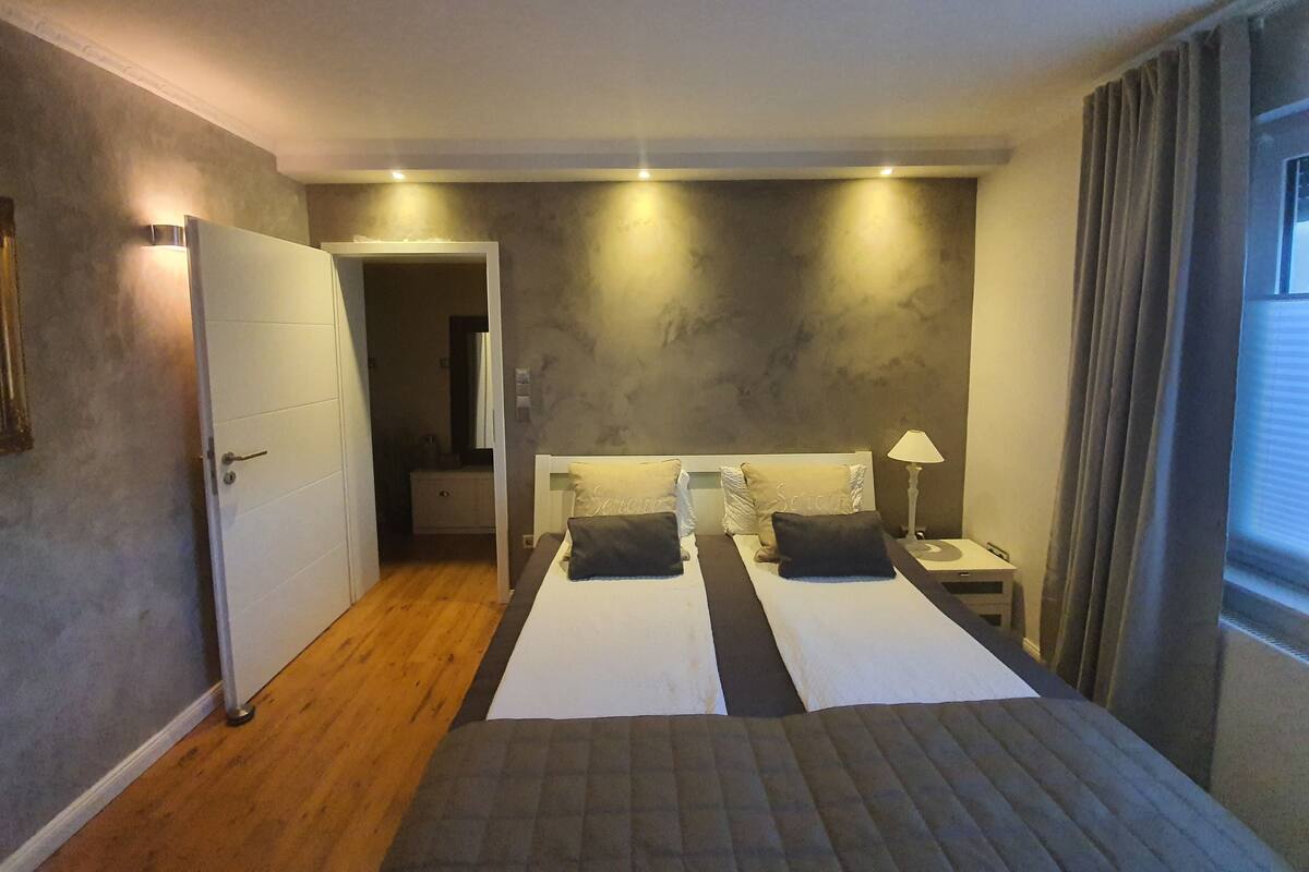 A hotel room with two beds, a bedside table with a lamp, a door leading to a bathroom, and a window with curtains.