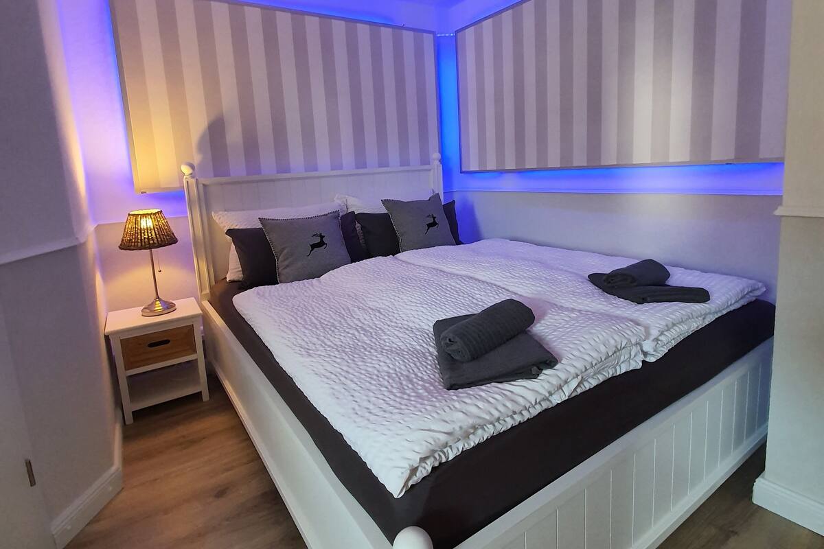 A neatly made bed with a white headboard and footboard, a bedside table with a lamp, and striped wall panels.