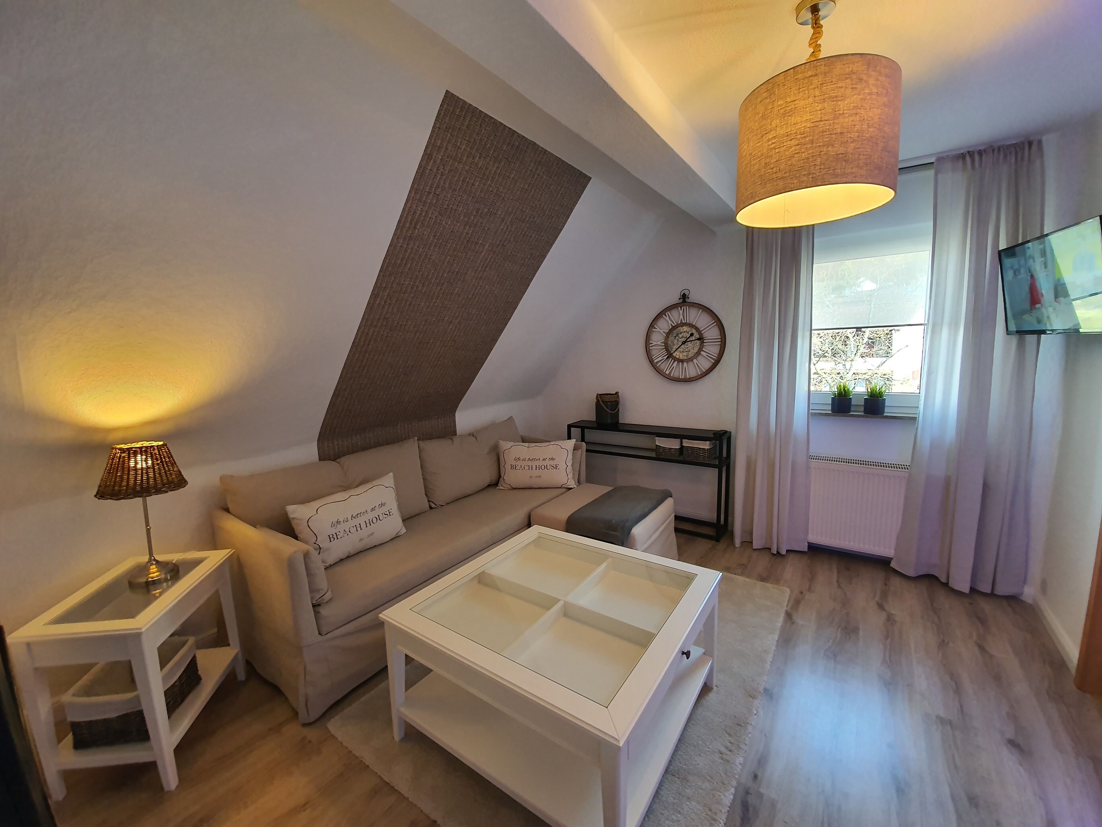 Un salon moderne comprenant un canapé, une table basse en verre, une table d’appoint, une lampe, une horloge murale, un téléviseur et une fenêtre avec des rideaux.