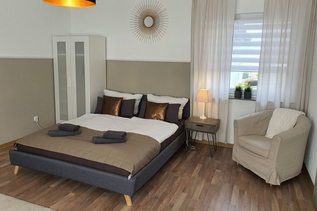 A modern hotel room with a large bed, a bedside table with a lamp, a chair, and a view of the outdoors through a window with sheer curtains.