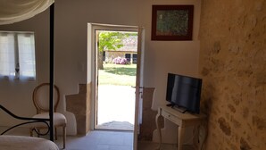 A room with a bed, a chair, a desk with a television, and a door leading to an outdoor area with a garden and a building. - Domaine de Manilau - B&B (Le Buisson-de-Cadouin)