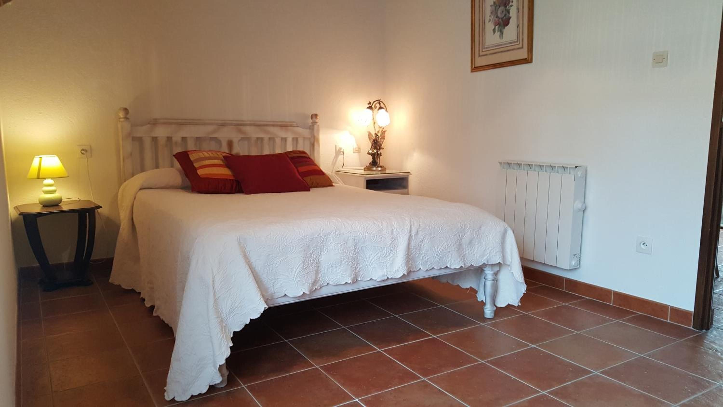 Un dormitorio con una cama, una mesita de noche con lámpara, un radiador y un cuadro en la pared.