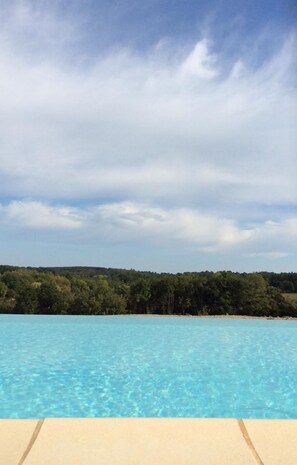 Une piscine d’eau bleue et limpide, d’où l’on peut admirer une colline boisée sous un ciel partiellement nuageux.