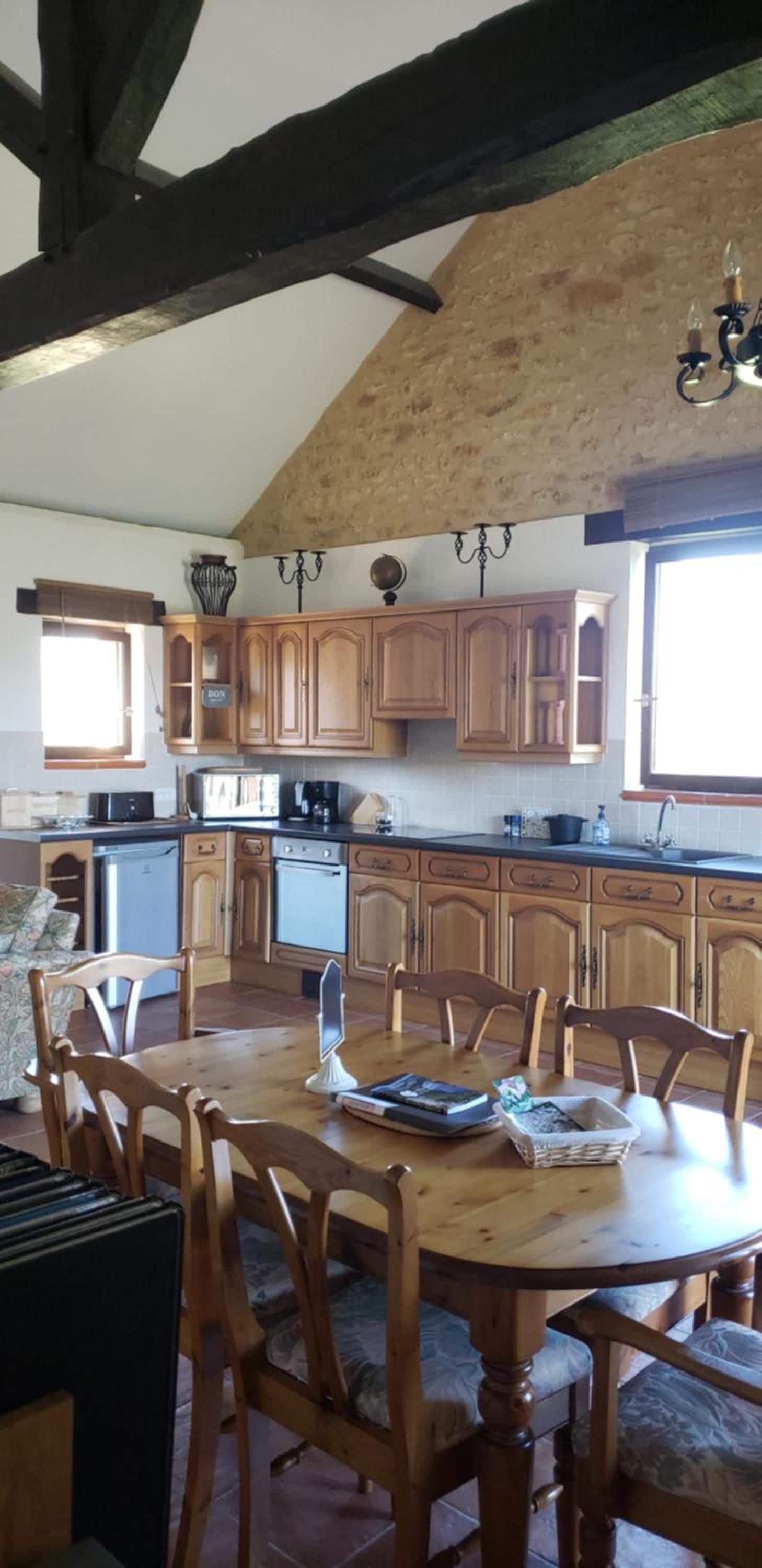 Een keuken met houten kastjes, een eettafel met stoelen, een koelkast en een spoelbak.