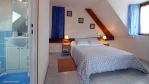 A bedroom with a bed, bedside tables, a nightstand, a window with curtains, and a bathroom with a sink and mirror. - Ferme Cidricole Hugues Desfrièches (Sainte-Marguerite-de-Carrouges)