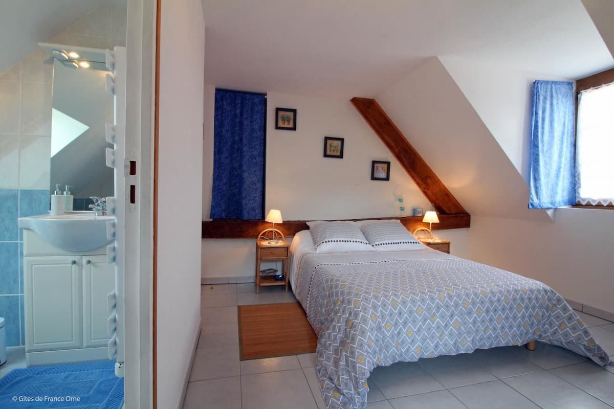 A bedroom with a bed, bedside tables, a nightstand, a window with curtains, and a bathroom with a sink and mirror.