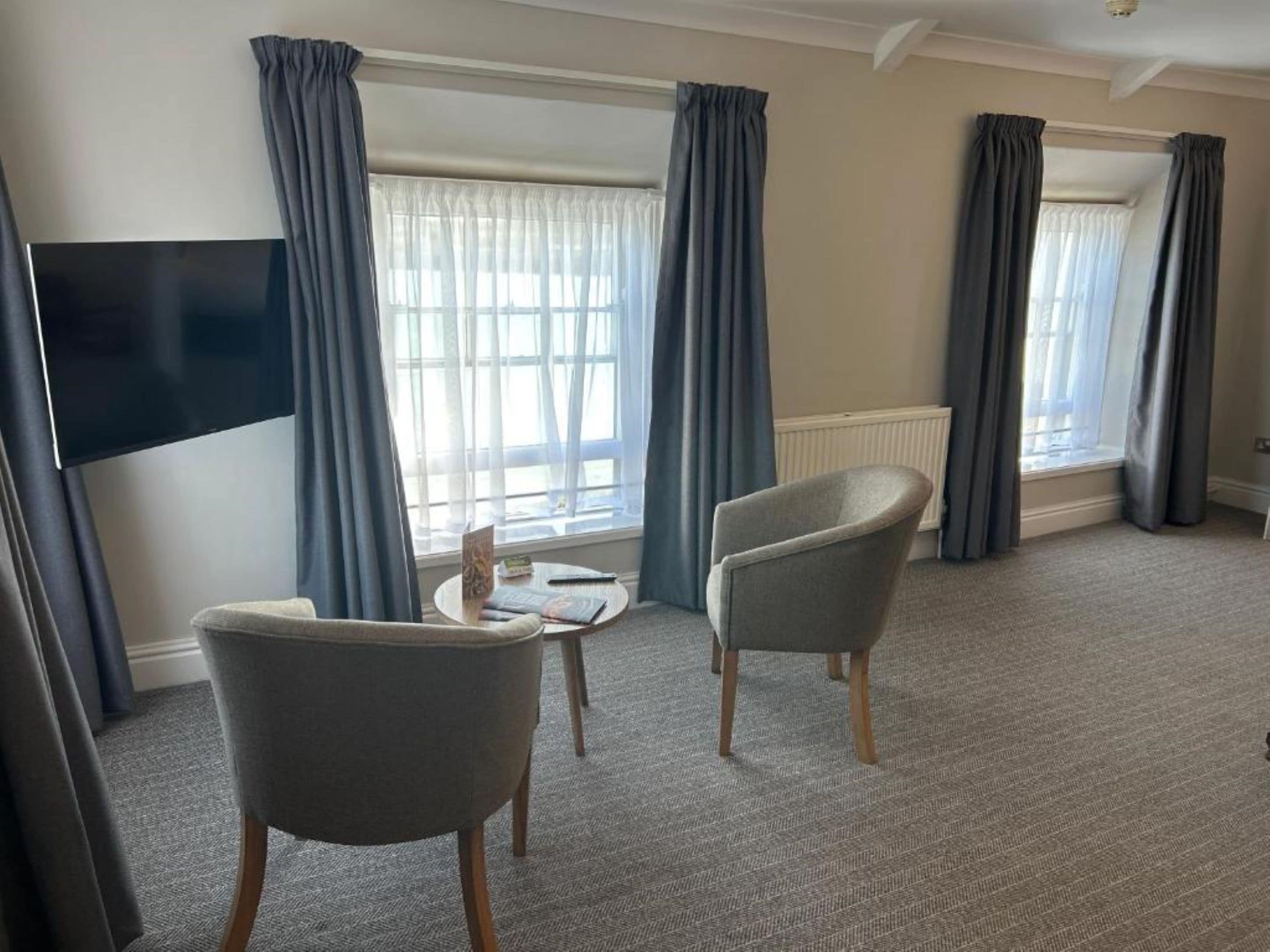Habitación de hotel con una televisión de pantalla plana grande, dos sillones grises, una mesa redonda pequeña y un ventanal con cortinas.