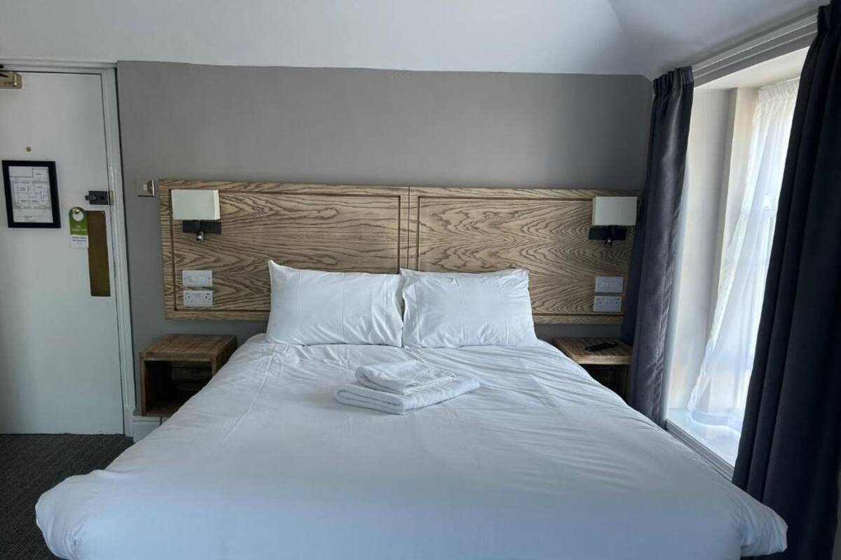 Cama bem arrumada com lençóis brancos, cabeceira de madeira, abajures e janela com cortinas.