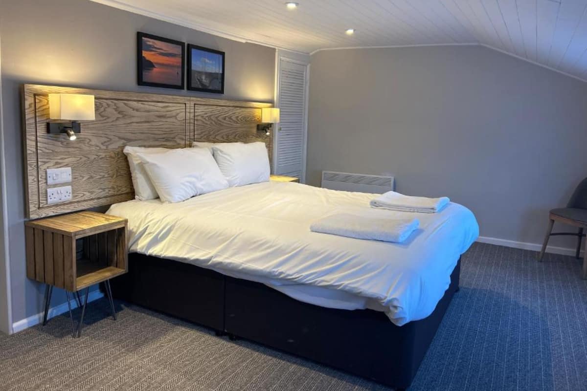 Quarto moderno com uma cama grande, cabeceira de madeira, abajures e uma pequena mesinha de cabeceira.