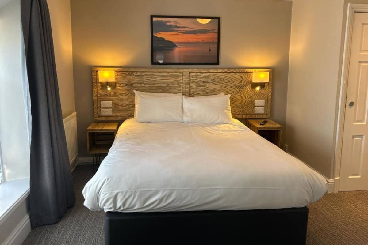 Cama bem arrumada em quarto de hotel, com cabeceira de madeira, duas luminárias de cabeceira e um quadro emoldurado acima da cama.