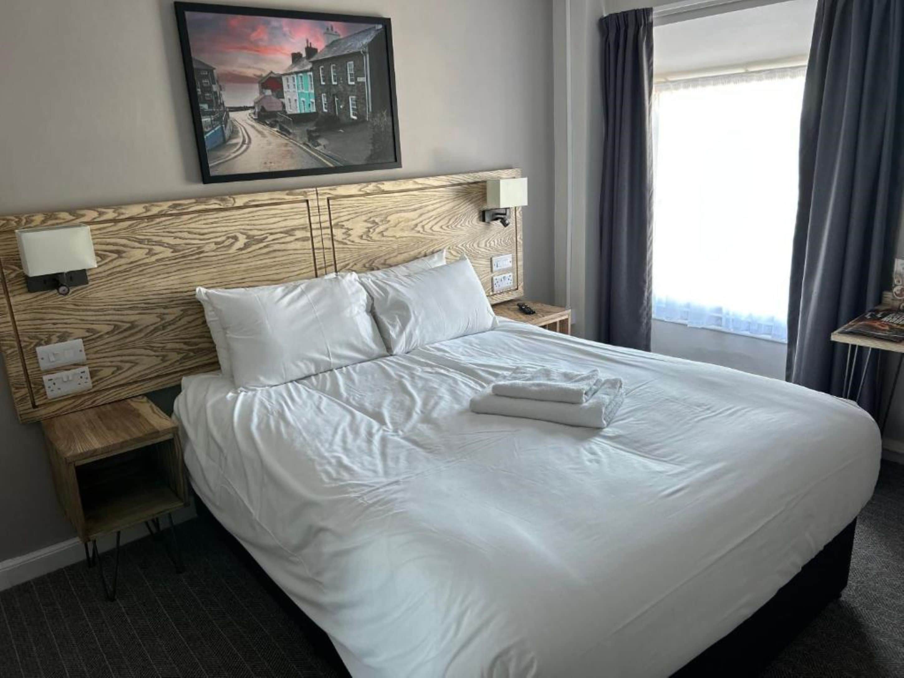 A neatly made bed with white linens, a headboard with a wood grain pattern, and a painting of a coastal scene above it.