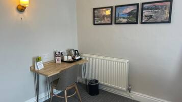 A small office space with a desk, chair, coffee maker, and a trash can. There are three framed pictures on the wall.
