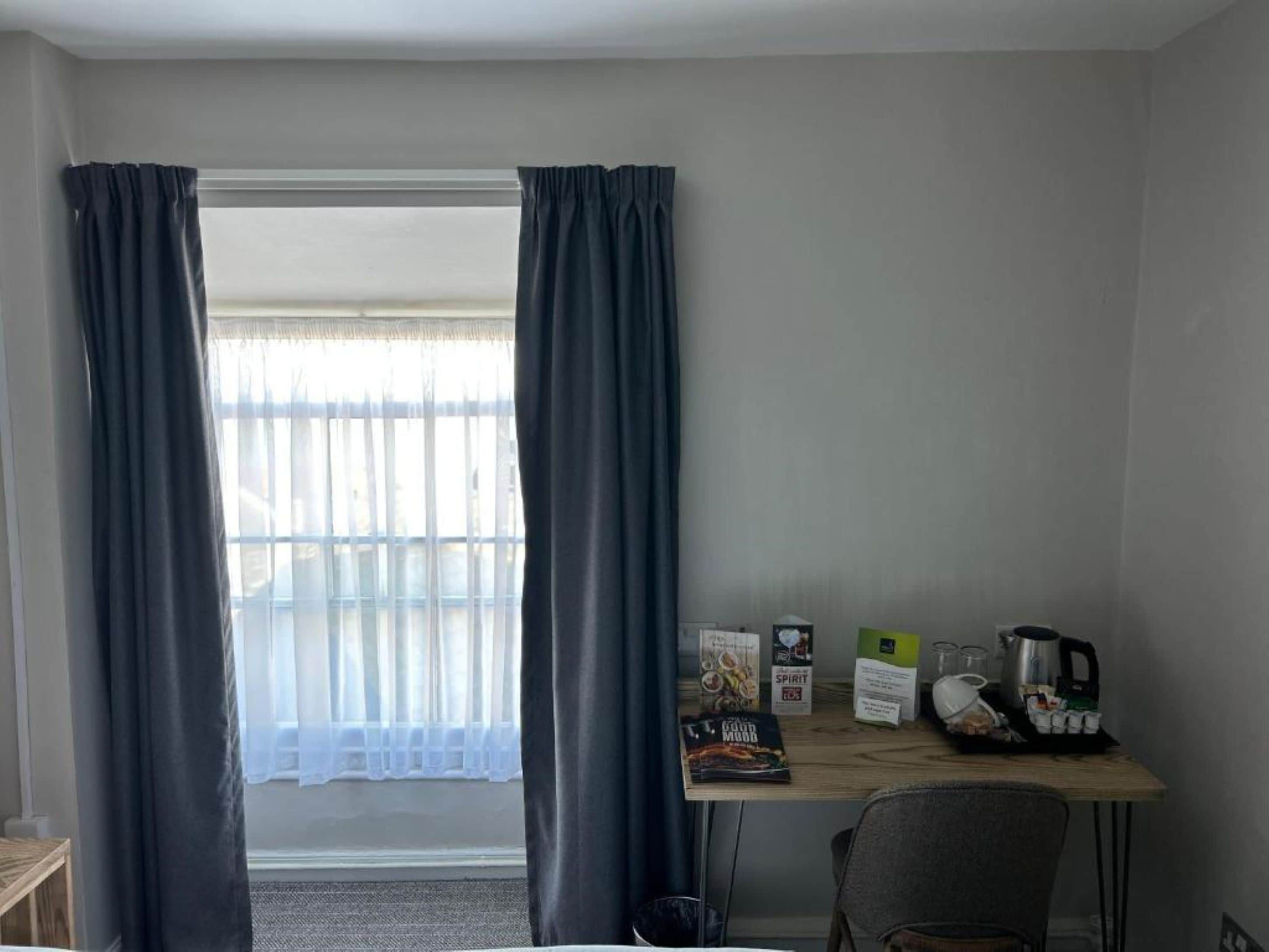 Una habitación con escritorio, una silla, un ventanal con cortinas y varios objetos sobre el escritorio, incluyendo una cafetera, una caja y una revista.