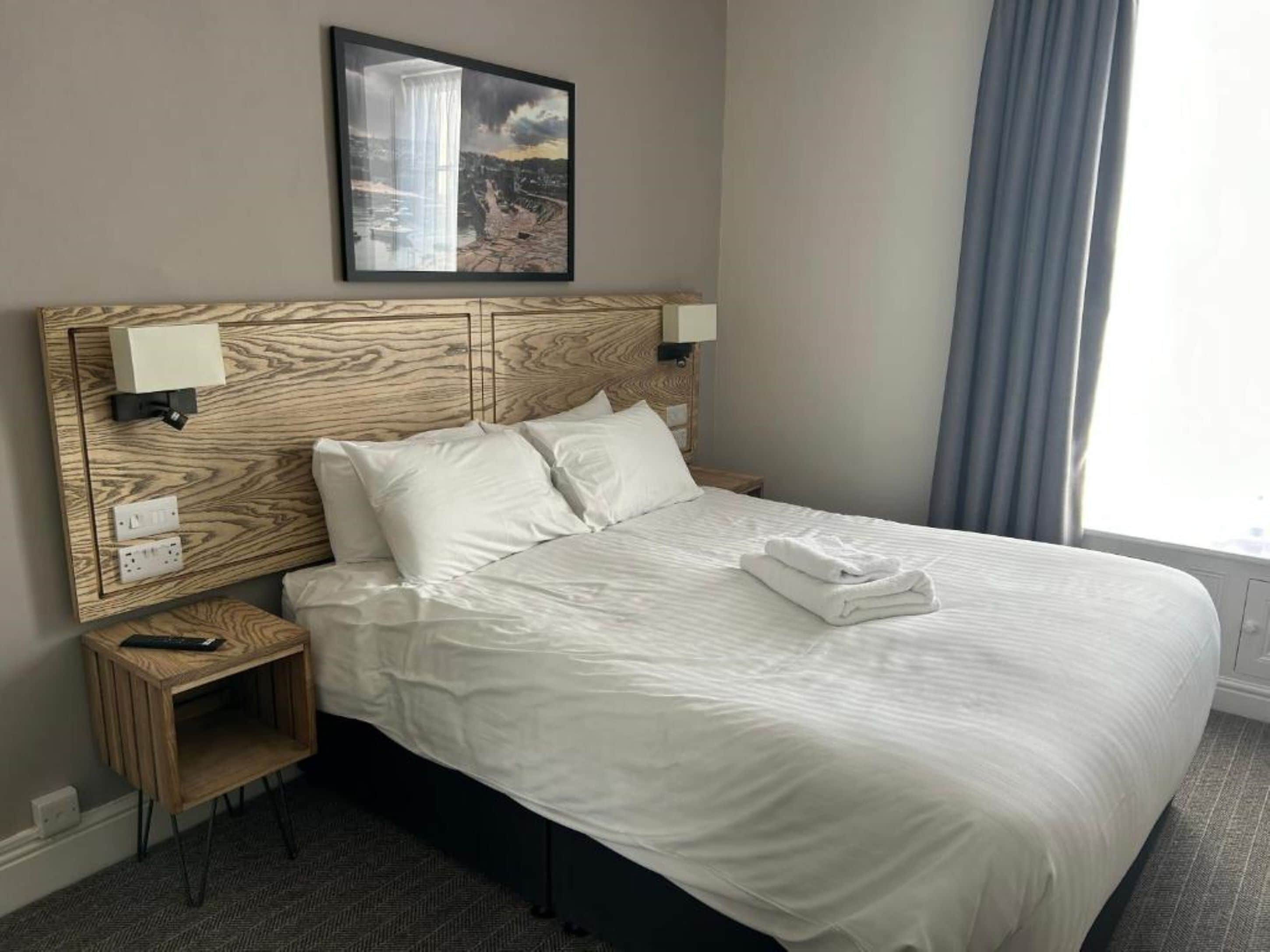 A neatly made bed with white linens and a folded towel, a wooden headboard with wall-mounted lights, a small bedside table with a remote control, and a framed picture above the bed.