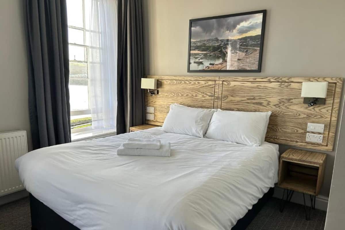 Quarto de hotel com uma cama grande, cabeceira com design de madeira, duas mesas de cabeceira com abajures e uma janela com cortinas.