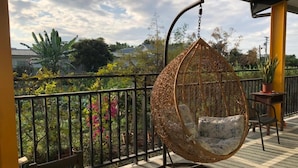 Une chaise suspendue sur un balcon, avec vue sur la verdure et une maison en arrière-plan.