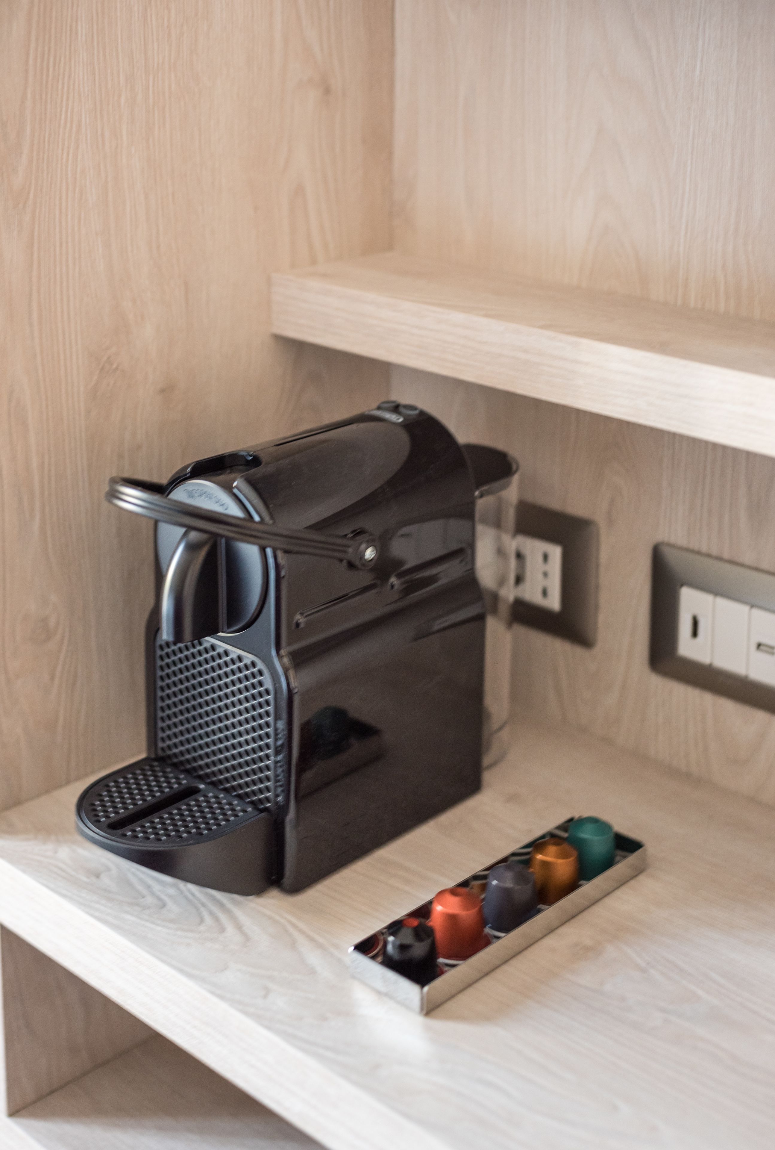 A compact coffee machine on a wooden shelf with a drawer containing colorful capsules.