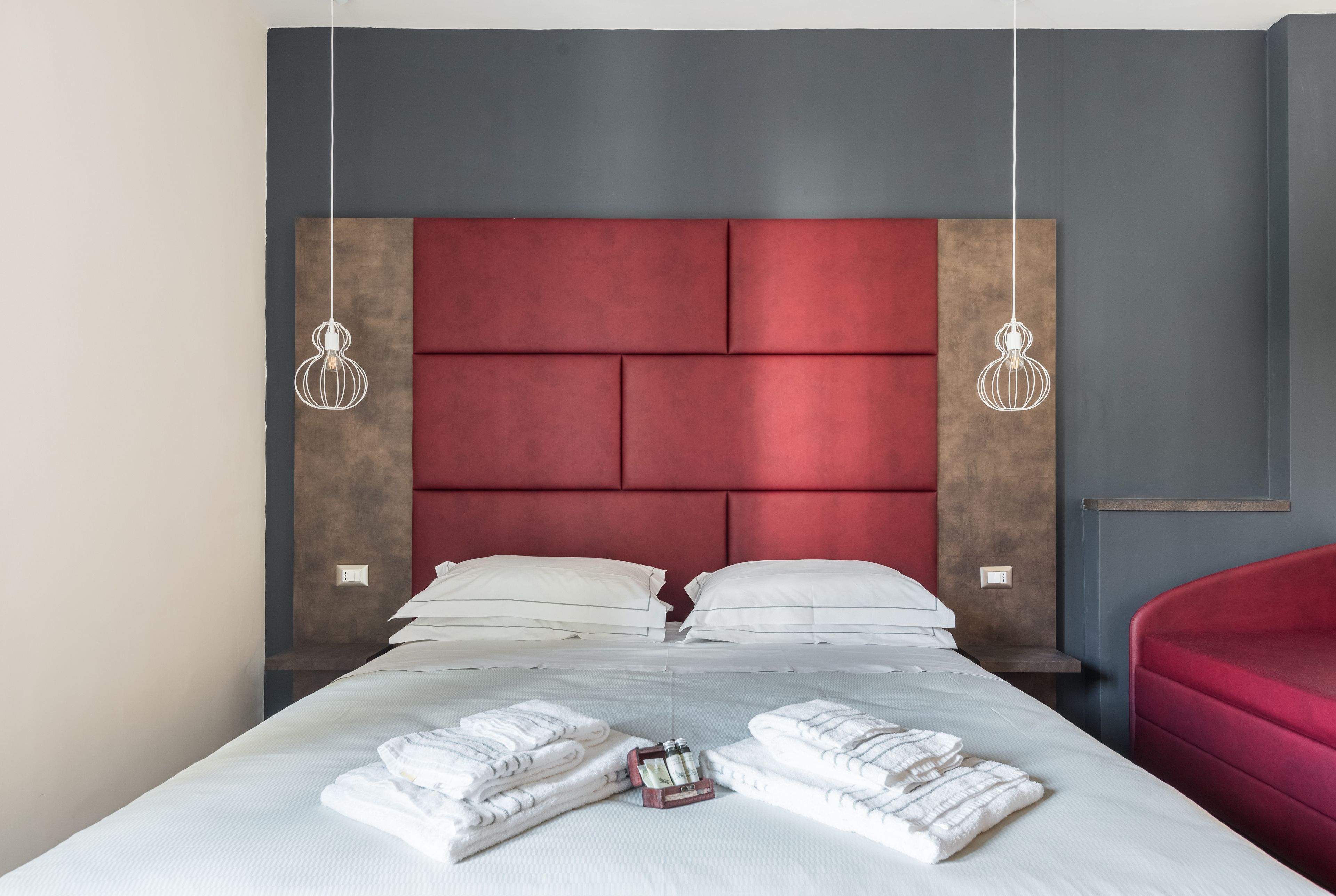 Habitación de hotel moderna con una cama grande, cabecera rojo y gris, ropa de cama blanca y dos lámparas colgantes.