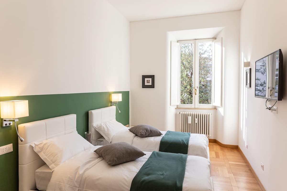 A hotel room with two beds, a window with a view, a wall-mounted TV, and a radiator.