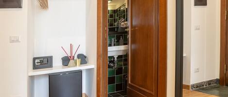 A hallway with a wooden floor, a white shelf with a telephone and decorative items, and a door leading to a room with a tiled floor.