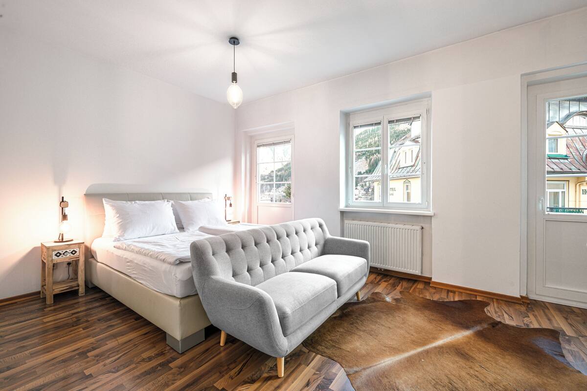 Ein Schlafzimmer mit einem Bett, einem grauen Sofa, Holzboden, einem Fenster und einer Heizung.